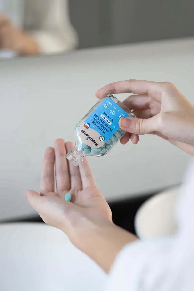 Een persoon giet een blauwe pil in een flesje met Tandpasta Tabs - Met Fluoride - Spearmint.